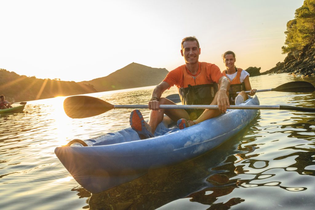 Candid portrait of Spanish friends in 30s and 40s pausing to smile at camera while paddling their tandem kayak off the Costa Brava.